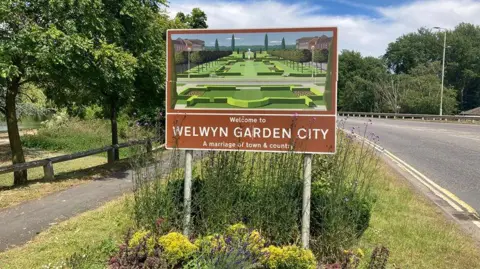A sign featuring an illustration of a formal garden in a brown frame with white lettering saying "Welcome to Welwyn Garden City". There is a road running past the sign. There are shrubs around the bottom of the sign and trees on either side of the picture.