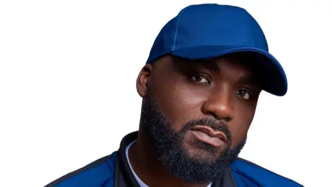 BBC DJ Ace wears a dark blue baseball cap and matching jacket in a head-and-shoulders portrait. He's got a neatly trimmed beard and a serious look on his face. He's standing in front of a bright white background, suggesting the shot was taken in a studio.