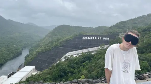 Ed Smith Ed Smith, blindfolded, standing beside view of dam in Thailand