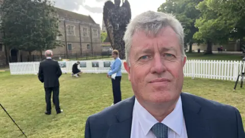 John Devine/BBC Jonathan James looks to the camera with the knife angel in the background - he wears a suit and checked tie