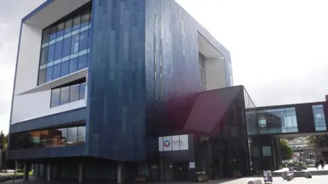 Geograph/Lewis Clarke Buckinghamshire New University campus in High Wycombe. Part of the building is curved with blue panels and houses the university's library. There is a walkway connecting campus buildings and a white sign which reads BUCKS NEW UNIVERSITY.