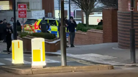 Ian Kershaw/BBC A GV of a police car outside a hospital with varilous passersby walking past 