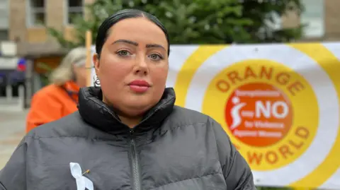 Suzie is wearing a dark padded jacket and stands outdoors in front of a large yellow and white banner reading “Orange World – Say No to Violence Against Women.” 