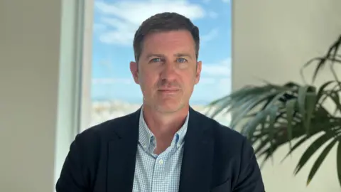 Photo of a man with short brown hair looking at the camera. He is wearing a navy blazer and a light blue and white checked shirt. 