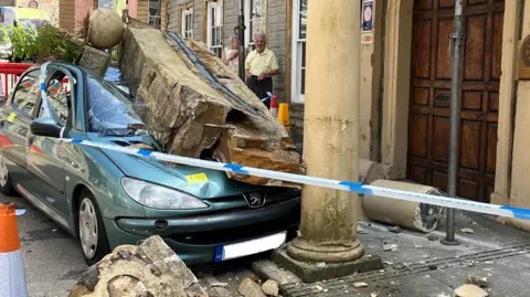 Car with smashed stones on top outside the pub. A large piece of masonry is resting on the bonnet and front window of the car