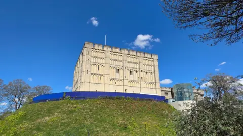 Norwich Castle, built of light-coloured stone, sits on top of a grassy mound. It has a flagpole but no flag flying