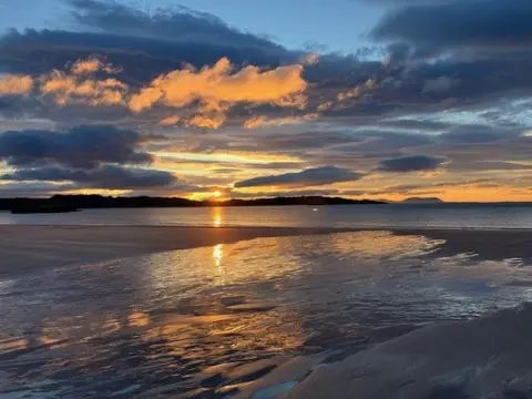 June Horn The sun sets above a beach, the sky is orange and blue
