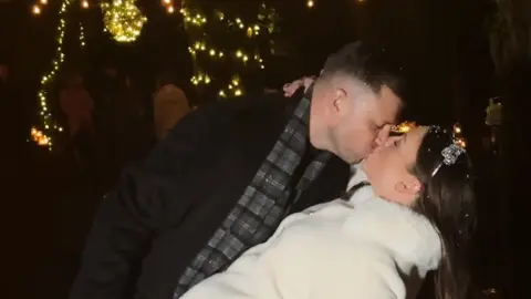 @reelallaboutit Michael and Sinead Lysaght kiss after marrying in a forest in December. She is wearing a white coat and silver hairband and he is wearing a dark coat with grey checked scarf. 