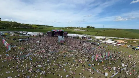 Aerial photograph of the festival's main stage area. Thousands of people are gathered on parkland.