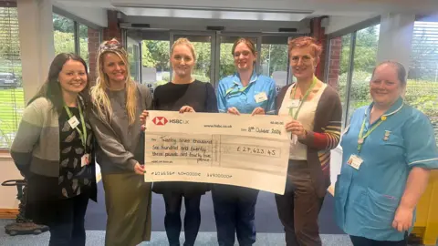 Prospect Hospice Several people including a couple in nurses' uniforms holding a large publicity style check in the entrance hall of a building, smiling and looking at the camera.