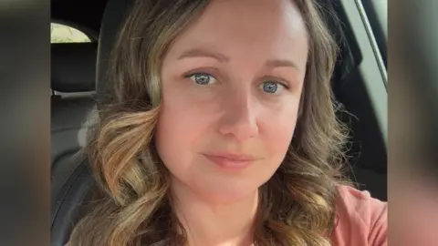 Alex Williams A woman with light brown curled hair. smiles, in a selfie. She is sitting in the front seat of a car, and wears a light pink top.