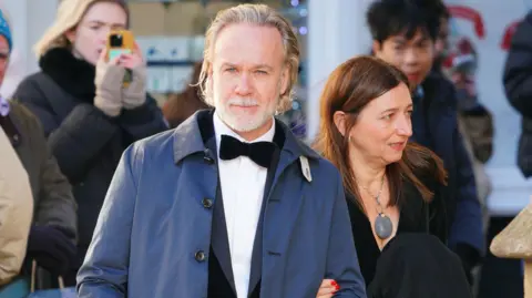 PA A man in a tuxedo and wearing a blue overcoat outside. His partner, a woman dressed in black, is holding his left arm.