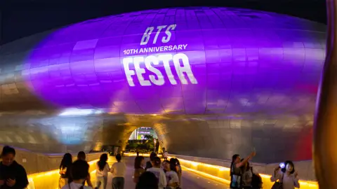 Getty Images People gather to celebrate the tenth anniversary of BTS at the Dongdaemun Plaza in Seoul, South Korea. Fans are taking pictures with a landmark building covered in purple, with a sign that reads "BTS 10th Anniversary".