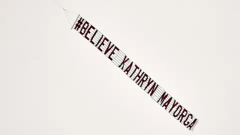 A banner reading "#Believe Kathryn Mayorga" flies over the stadium