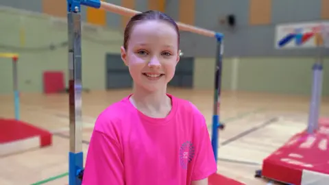 Maisie. She has brown hair tied back, wearing a pink t-shirt and smiling at the camera. Gymnastic bars are behind her.