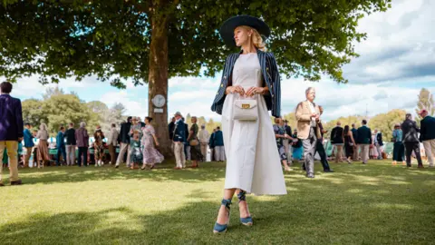 Henley Royal Regatta / Row360 A woman dressed in a striped rowing blazer over a white dress is posing for a picture. There are many other people behind her under a big tree. 