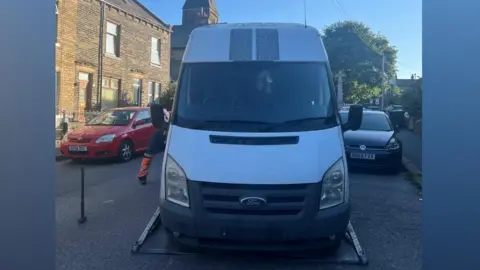 Front view of a white Ford Transit-style van parked on a residential street and positioned on the platform of a recovery truck. Brick terraced houses line the street on the left, with several parked cars visible on both sides, including a red car and a dark blue car. A church tower rises in the background behind the van. Bright sunlight casts strong shadows, and part of a person wearing high-visibility orange trousers is visible beside the recovery vehicle. The van's windscreen is darkly tinted, and patterned