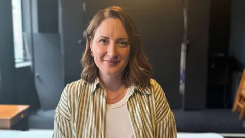 BBC A woman with shoulder length brown hair. She is wearing a green and white striped shirt opened over a white vest top. She has a thin gold necklace around her neck and one gold earring can been seen with her hair tucked behind her ear. She is in front of a black background with a wooden beside drawer to her right. 