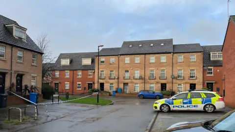 A police car parked in a residential street, opposite a house with police tape blocking off the outside.