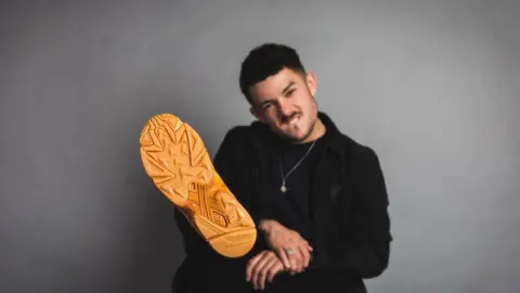 Sorley has short dark hair with dark stubble and is biting his lips. He is wearing dark clothing with a silver chain over his top. His leg is raised up with the orange sole of his shoe facing the camera