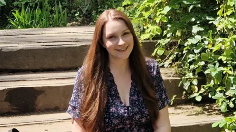 Megan McCley Megan McCley sitting in a purple and pink floral dress smiling at the camera in sunshine. The backdrop is wooden steps in what appears to be a garden, with foliage to her left.