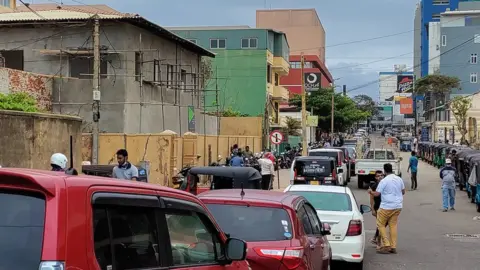 A street sloping upwards is filled with cars in single file stretching as far as the eye can see.