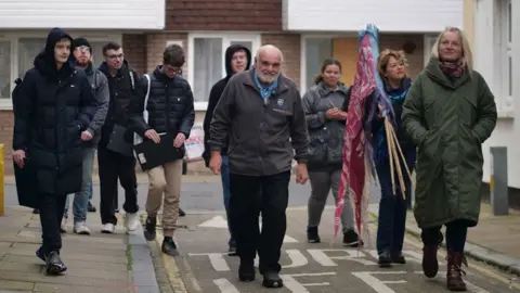 Marley Karazimba A group of adults in winter clothes walking along a street in Harwich. They are led by a man in a grey jacket with white hair and beard. A woman on teh right is holding a number of flags on poles over her shoulder