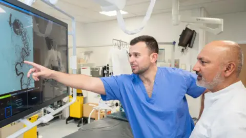 Leeds Teaching Hospitals A man in blue scrubs points at a tangle of lines on a computer screen while a man with short white beard and a white shirt watches
