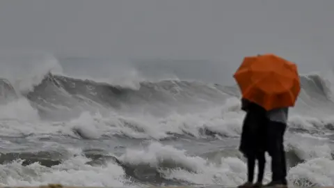 Getty Images A couple stands holding an umbrella amid gusty winds as dark clouds loom over Marina beach in Chennai on December 8, 2022, ahead of Cyclone Mandous forecasted landfall in north Tamil Nadu-south Andhra Pradesh coasts. (Photo by Arun SANKAR / AFP) (Photo by ARUN SANKAR/AFP via Getty Images)