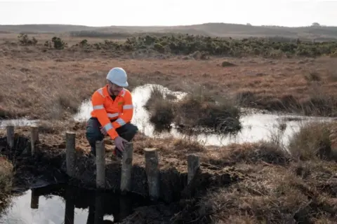 Sophie Bolesworth Man in orange hi-vis and white hard hat next to one of the leaky dams