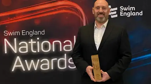 Ben Negus wearing a black suit and a white shirt- holding a brown trophy - standing in front of a black and red national swim awards poster