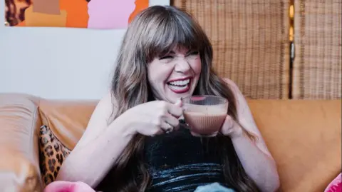 Ciara Hillyer Briony May Williams holding a cup of coffee while sitting on a leather sofa. She has long dark brown hair and is wearing red lipstick. 
