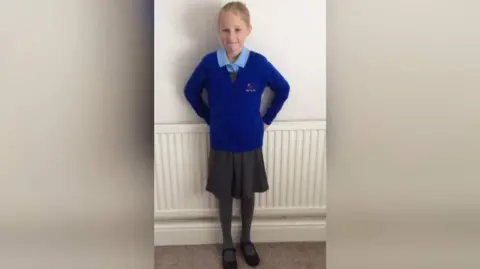 Rachel Cowley, una niña, con cabello rubio con un uniforme escolar azul, una falda escolar y zapatos de escuela negros. Ella está parada frente a un radiador apoyado en una pared blanca.