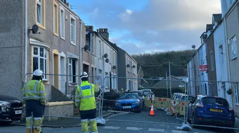 Officers in high-vis jackets stand on a street which is partly fenced off. A house has visible damage to its first flood and roof and a car parked outside also has its roof and windscreen smashed.
