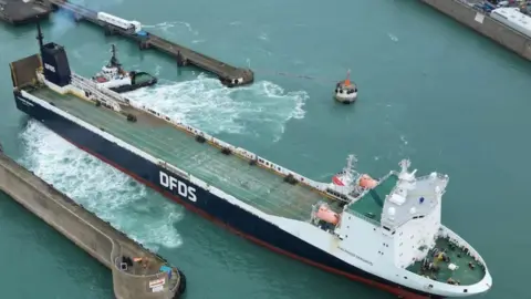 A image of a DFDS ferry docked up at a stopping spot. Pictured is a long ferry which has the letters DFDS written on its side. It has a white end and a navy body.