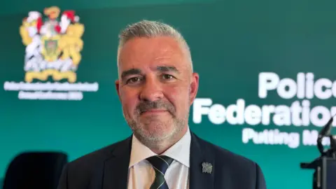 Liam Kelly, smiling with his mouth closed, stands in front of what seems to be la projector screen. On it is the emerald green branding of the Police Federation of Northern Ireland, with its insignia in the top left corner and white writing in the centre. Liam is wearing a dark suit and has shirt grey hair and a light stubble of the same colour.