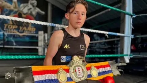 EMMA BAUGH/BBC Jerry-Mike wearing red boxing gloves and sitting on the boxing ring with his championship belt.