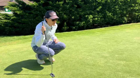 Michelle Lau is squatting onto her ankles to get a better look at  where her golf ball is. She is also holding her golf club. She is wearing sunglasses, a grey cap, white top and purple trousers.