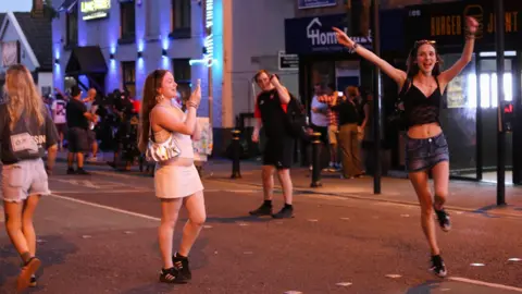 Emma Gibbs A woman in a white dress and black trainers films another woman, in a black top, denim skirt and black trainers, on her phone as she dances in the street after Oasis at Heaton Park