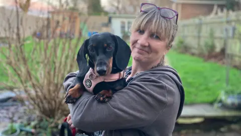 Ellen Ashwood holds Pepper, a black sausage dog, in her arms, and both look at the camera. Pepper has brown paws and is wearing a pale pink harness, in the background you can see a garden with lawn and some bare shrubs.