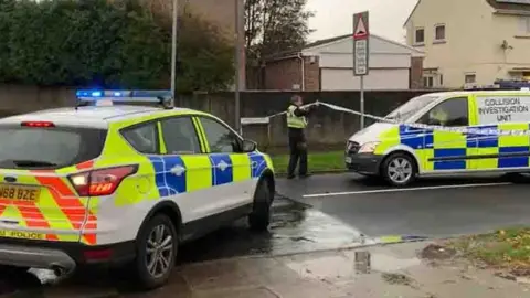 A police officer lifts a police cordon. To the left of the picture is a police car and on the right is a collision investigation unit van