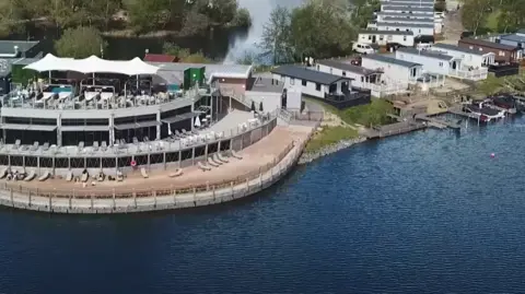 Away Resorts An aerial image of Tattershall Lakes Country Park shows the shore of a lake with several long, thin static caravans on the shore and a couple of small boats moored. There is also a large communal area and beach with sun loungers and a large white umbrella offering some shade.