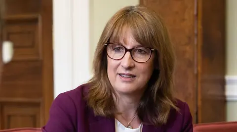 A close up image of Rachel Reeves wearing glasses and a purple suit while sitting on a sofa as she talks to Sir Charles Roxburgh (out of frame) in London on Monday.