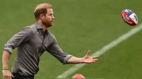 Prince Harry, in a grey shirt, plays with an AFL football