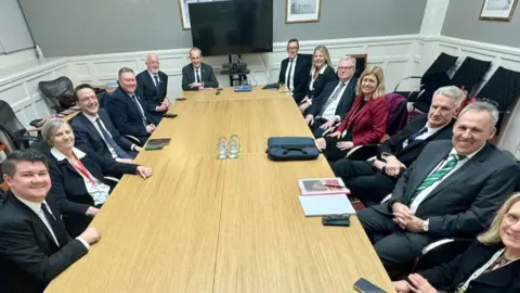 Deputy Lyndon Farnham's ministerial team are sat around a long light brown table. They are in a meeting room with a TV at the end of the table. There are thirteen ministers around the table including the Chief Minister. 