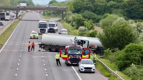 An empty motorway with a truck across the road and into the barrier and hedge. There is a fire truck and emergency services in high vis jackets.