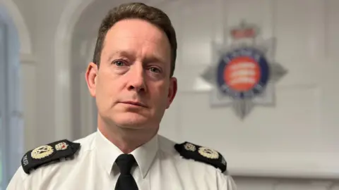 Ben-Julian Harrington, who has short brown hair, staring at the camera with a serious expression. He is wearing a white shirt with a black tie and black epaulettes, which have a golden crest and crown on them. In the background is the blue, red and silver Essex Police crest.