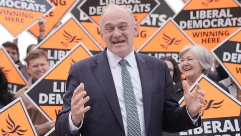 Liberal Democrat leader Sir Ed Davey stands in front of a group of supporters in Guildford, Surrey, holding orange Liberal Democrat signs.