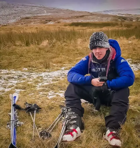 Chris Morris Chris sitting next to his skis in some grass with snow on a mountain in the background