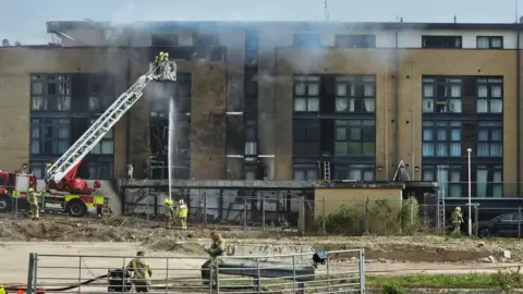 Michael Keohan/BBC Firefighters extinguish a fire in a block of flats. Smoke is coming off a brown black of flats, with parts of the building now black from where fire has damaged it.